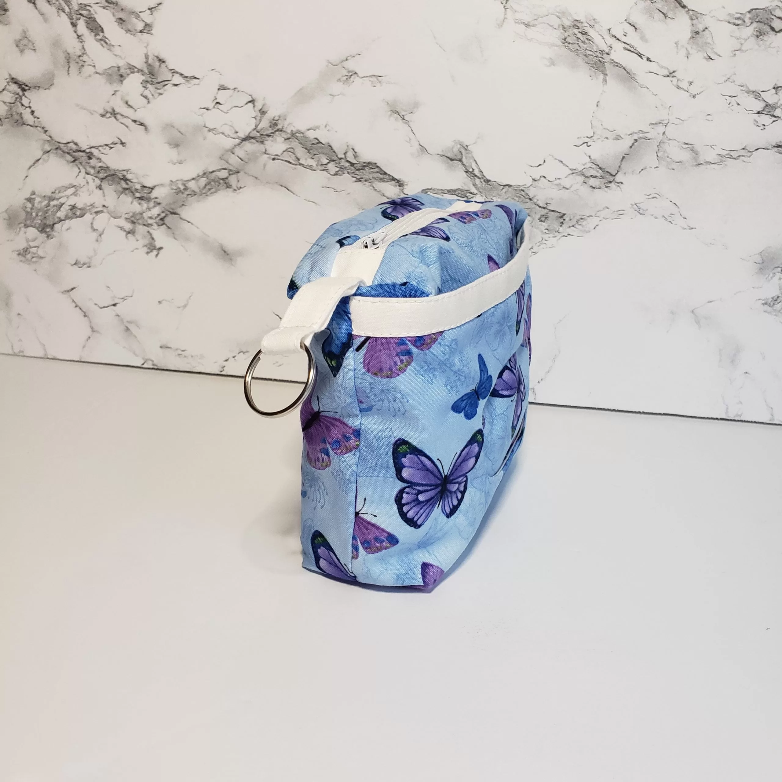 Small blue butterfly pattern accessory bag with front window pocket for makeup remover wipes. Fully lined interior. Zip closure with attached keyring. Measures 6 inches wide. Window pocket allows easy access to a packet of wipes (not included).