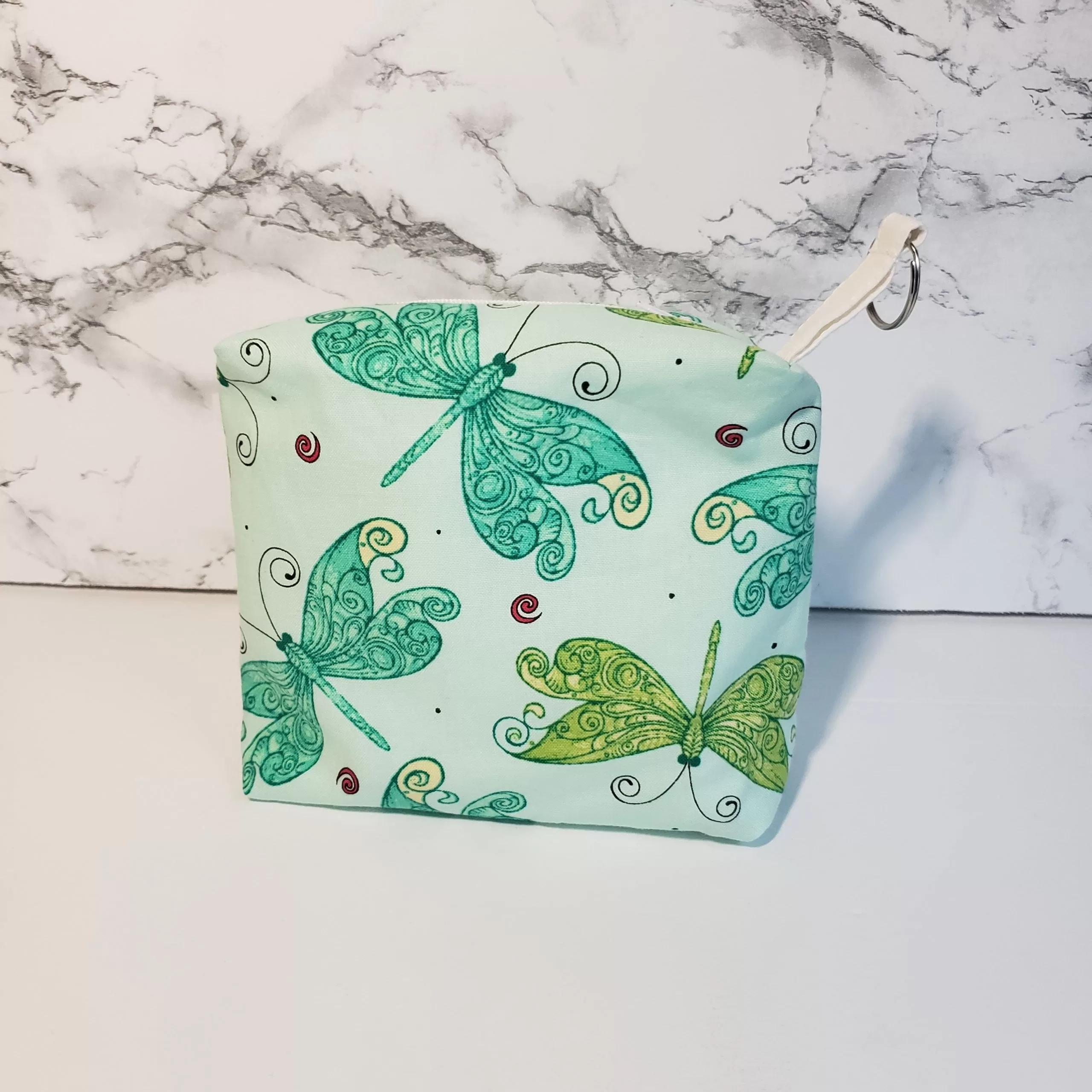 Small green dragonfly pattern accessory bag with front window pocket for makeup remover wipes. Fully lined interior. Zip closure with attached keyring. Measures 6 inches wide. Window pocket allows easy access to a packet of wipes (not included).