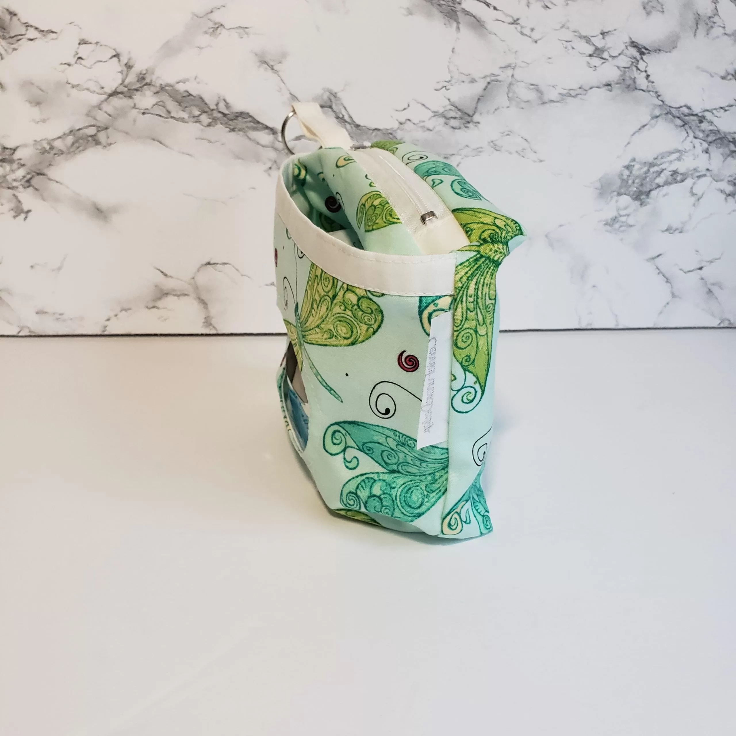 Small green dragonfly pattern accessory bag with front window pocket for makeup remover wipes. Fully lined interior. Zip closure with attached keyring. Measures 6 inches wide. Window pocket allows easy access to a packet of wipes (not included).