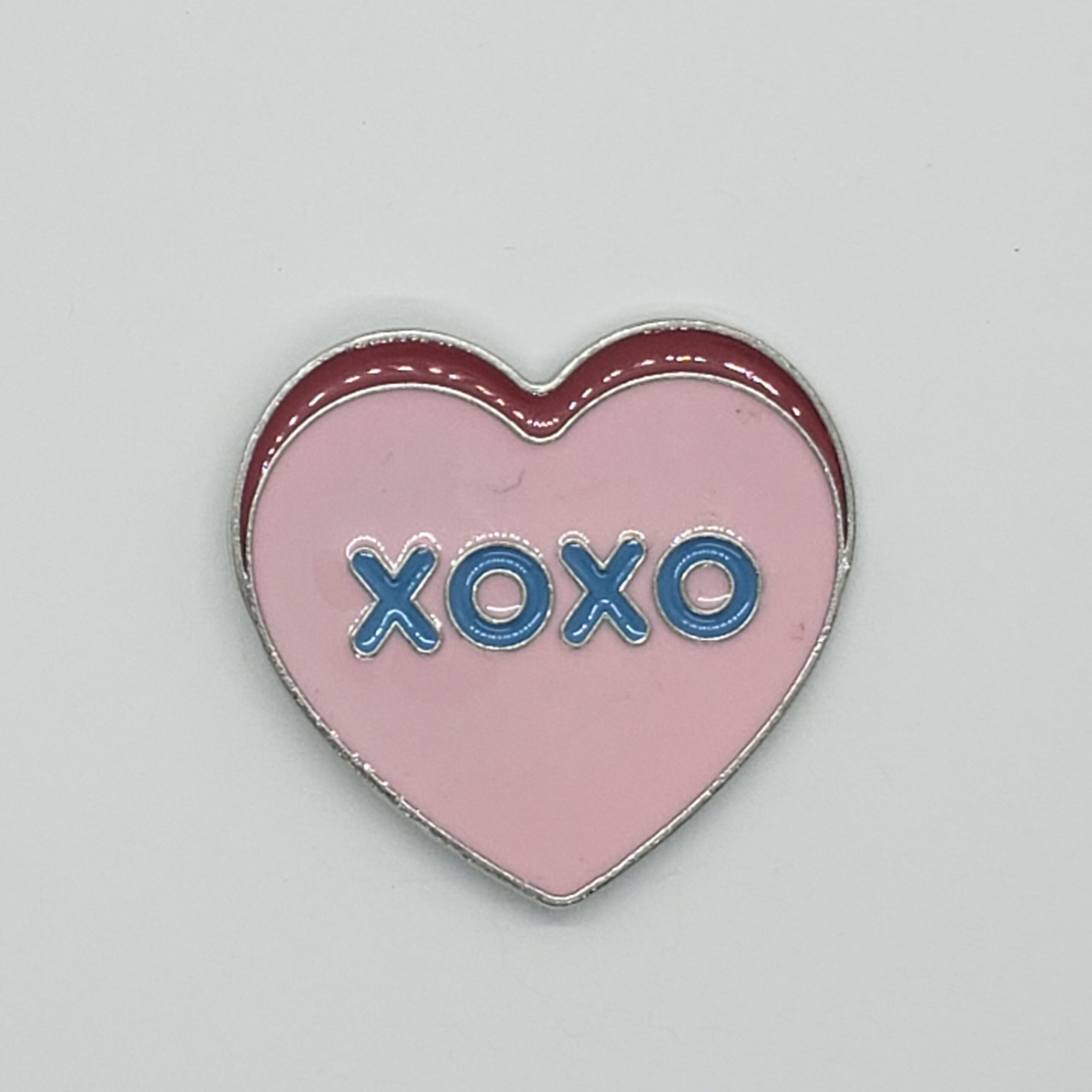 Large heart shaped enamel pin. Pink, with the phrase "XOXO" in blue text